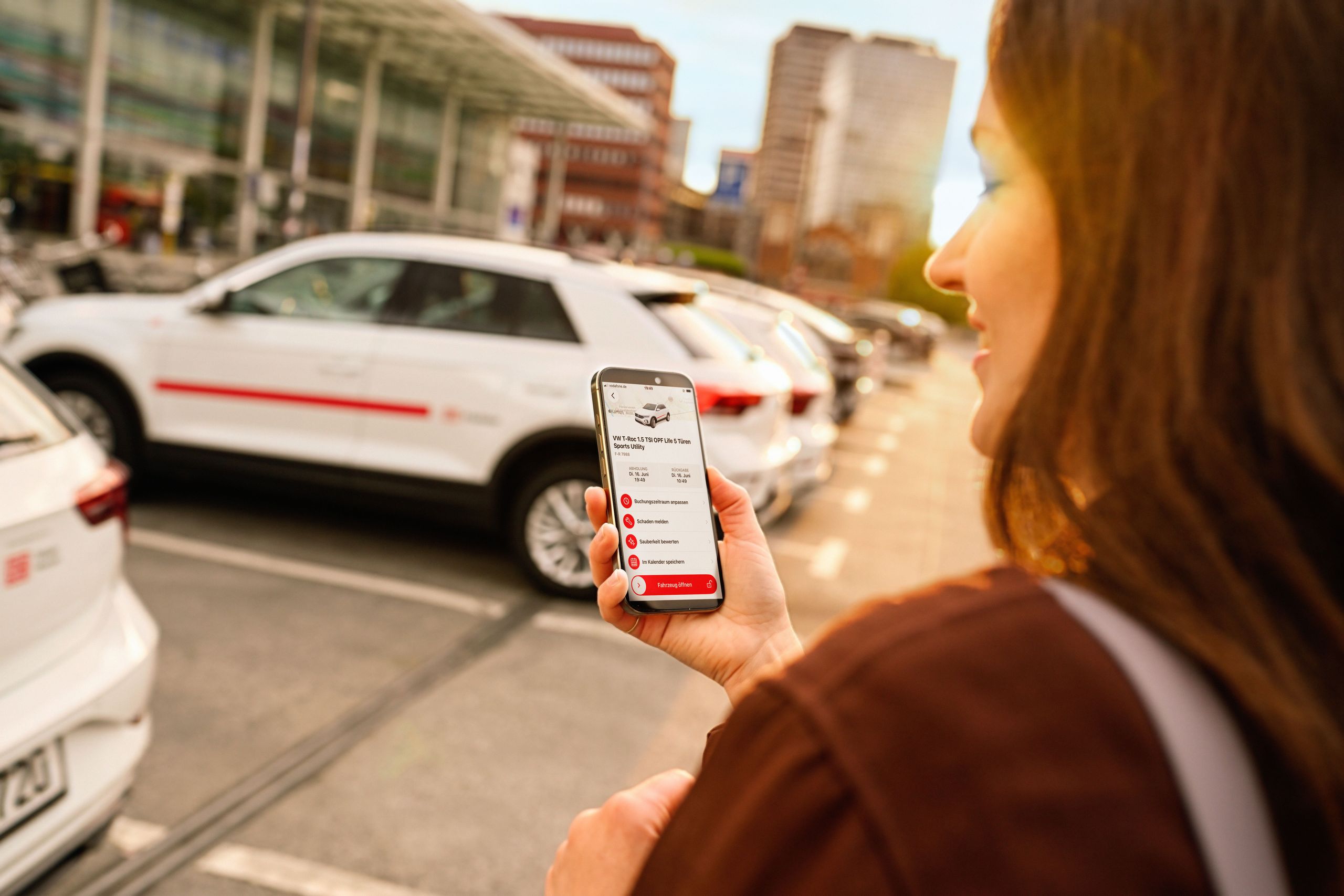 Flinkster-Kunde vor einem weißen Flinkster-Auto, schaut auf sein Smartphone, auf dem die Flinkster-App geöffnet ist.