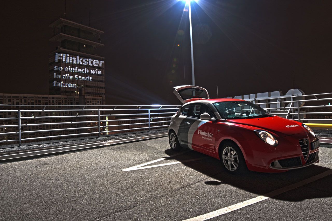 Flinkster-Auto mit offenem Kofferraum bei Nacht auf einem Parkplatz, welches die Botschaft 'Flinkster so einfach wie in die Stadt fahren' auf eine dunkle Hauswand projiziert.