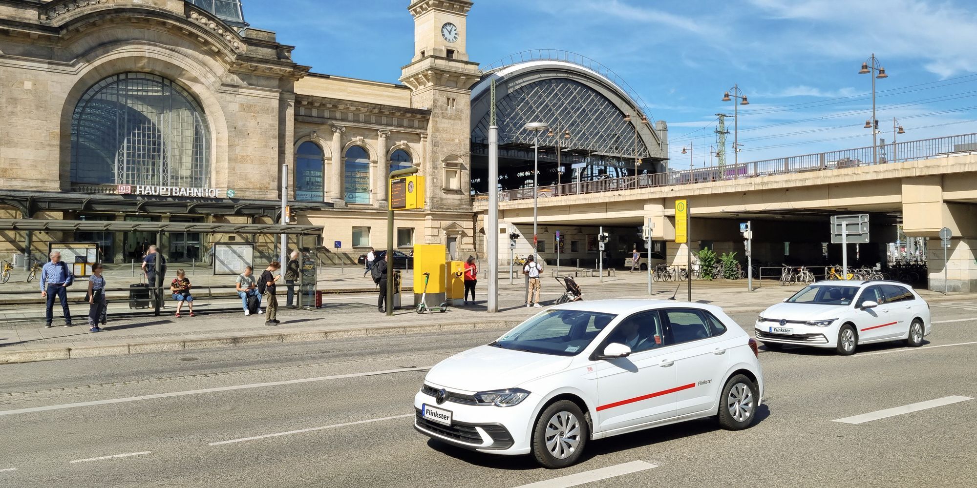 Zwei weiße Flinkster-Autos vor einem historischen Bahnhofsgebäude mit Uhrturm und DB-Logo an einem sonnigen Tag.