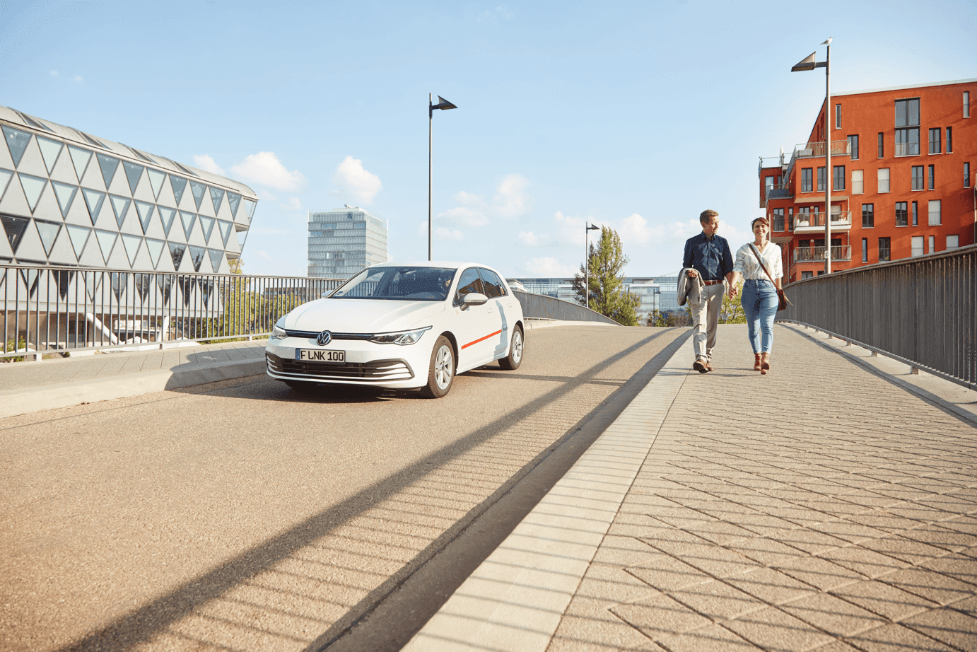 Bild auf einer Brücke mit zwei spazierenden Personen (Mann und Frau) und einem weißen Flinkster Fahrzeug, was an Ihnen vorbei fährt.
