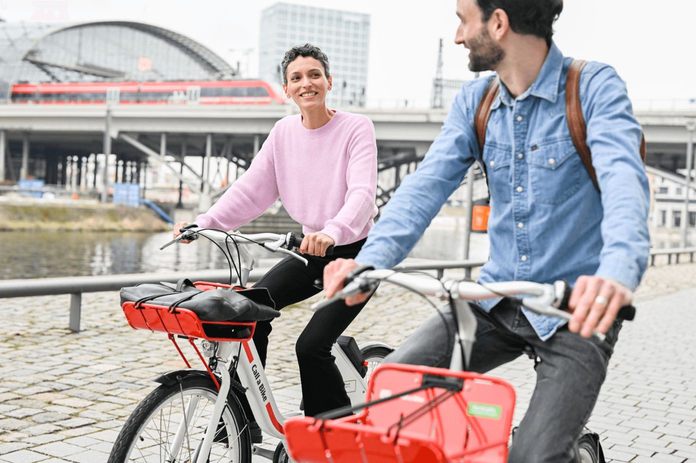 Zwei Personen fahren auf Call a Bike Fahrrädern an einem Fluss entlang, mit dem Berliner Hauptbahnhof und einem Zug im Hintergrund.