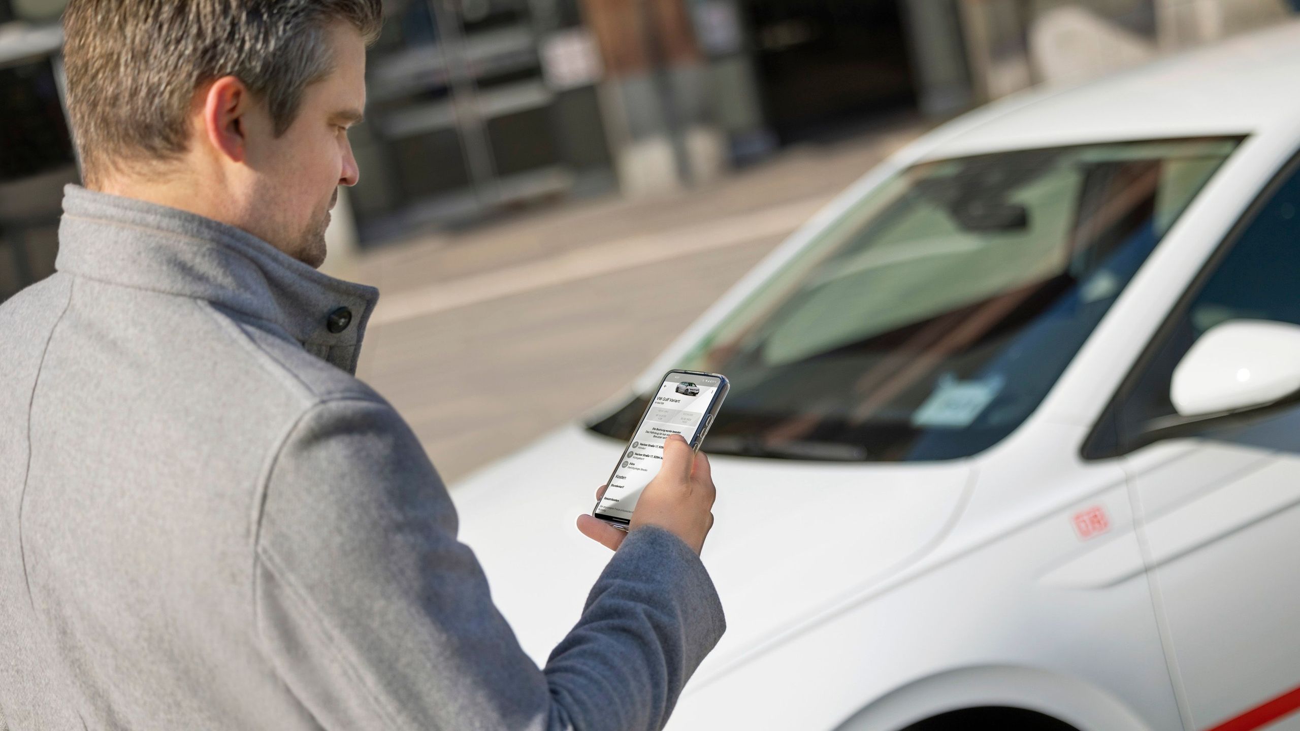 Flinkster customer in a gray coat in front of a white Flinkster car, looking at his smartphone on which the Flinkster app is open.