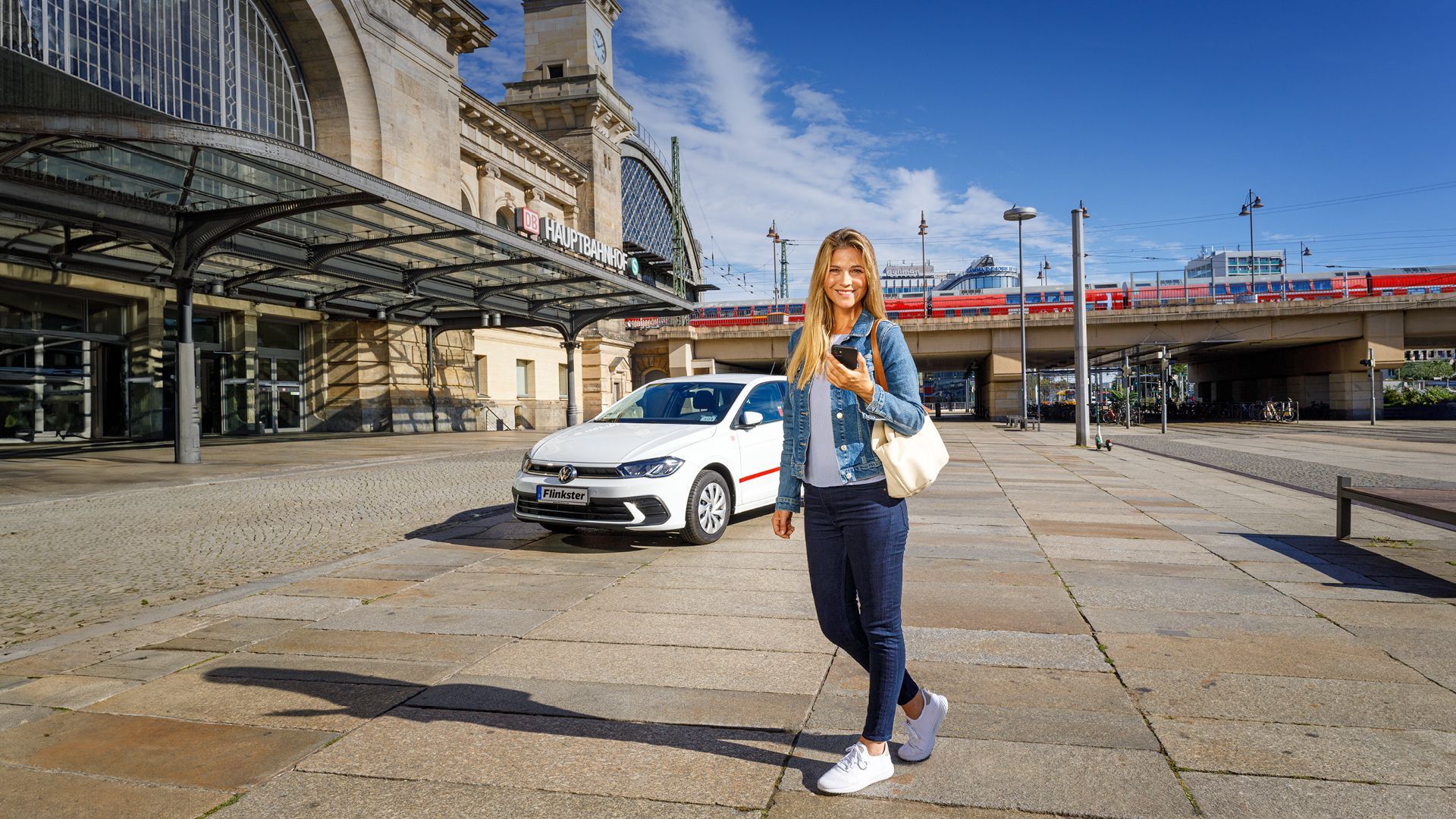Frau mit Smartphone und Umhängetasche am Bahnhof mit Flinkster Fahrzeug im Hintergrund.