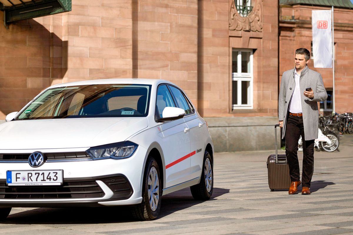 Ein Mann mit Mantel und Rollkoffer geht auf ein weißes Flinkster-Auto zu, das vor einem historischen Bahnhofsgebäude mit DB-Logo geparkt ist.