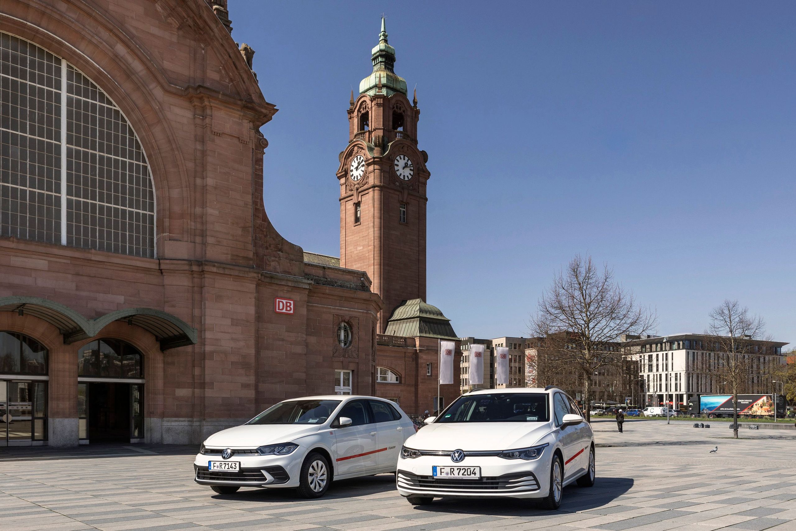Zwei weiße Flinkster-Fahrzeuge, ein VW Polo und ein VW Golf Variant, fahren vor dem Hauptbahnhof vorbei. Im Hintergrund stehen wartende Personen an einer Bushaltestelle.