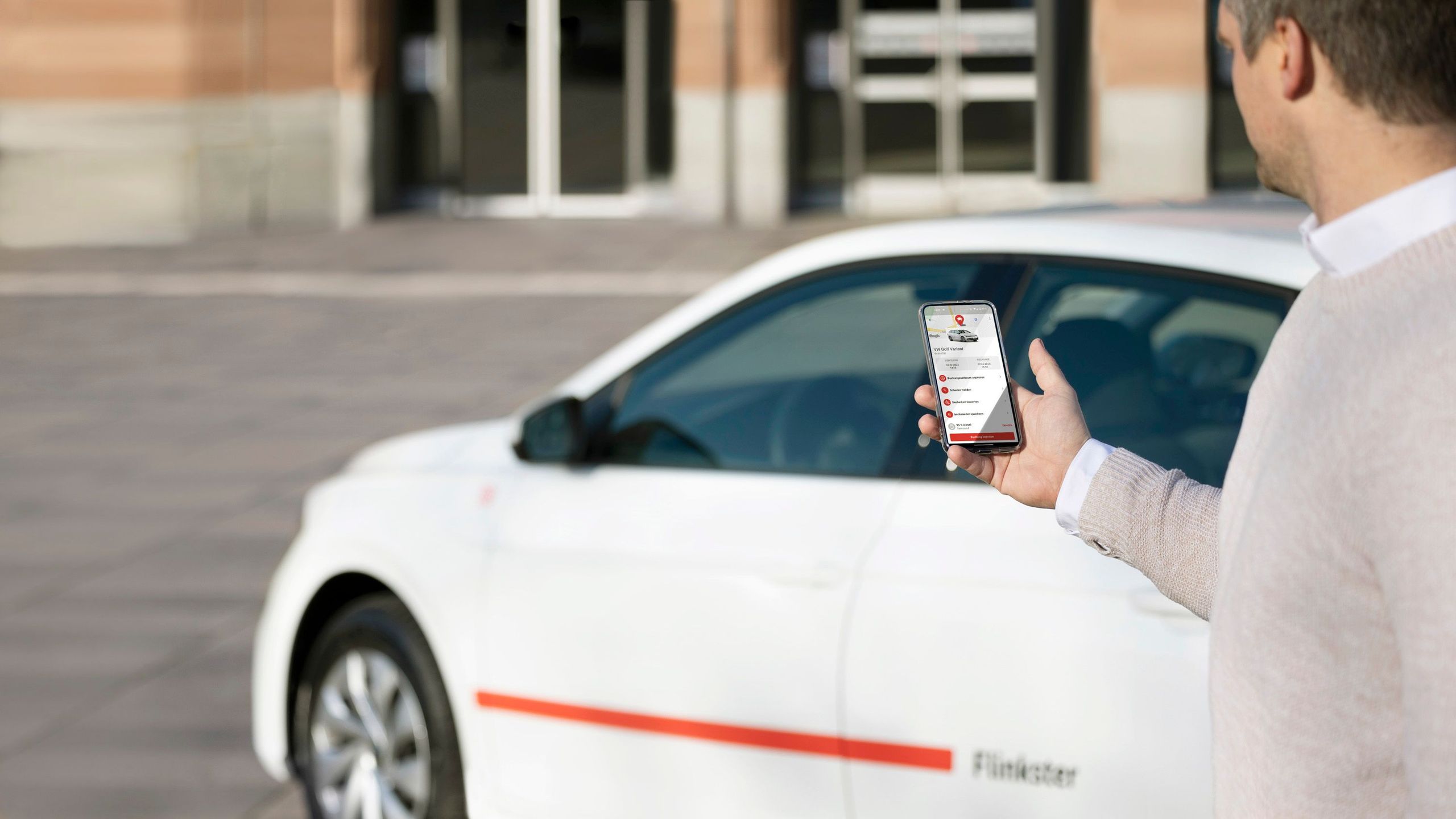 Flinkster-Kunde in einem grauen Mantel vor einem weißen Flinkster-Auto, schaut auf sein Smartphone, auf dem die Flinkster-App geöffnet ist.