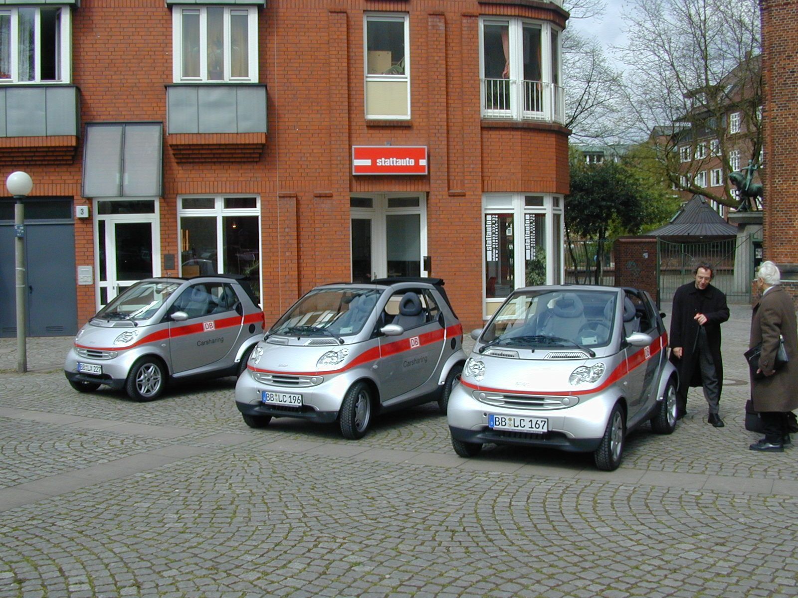 Drei silberne DB Carsharing-Autos und zwei Menschen vor einem roten Backsteingebäude.