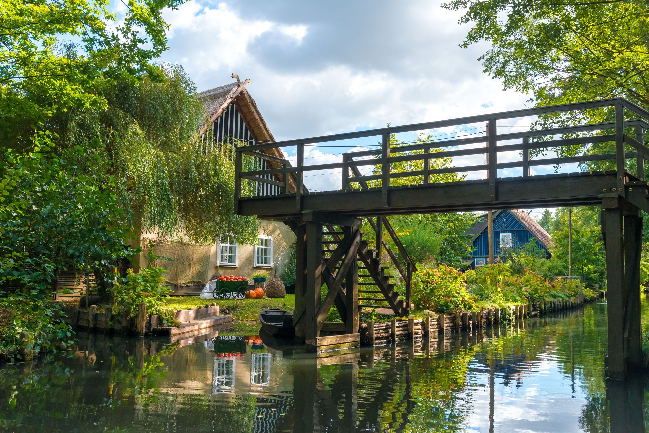 Holzbrücke über einen stillen Kanal im Spreewald, umgeben von üppigem Grün und einem reetgedeckten Haus im Hintergrund, das eine idyllische ländliche Szene darstellt. copyright Foto: istock.com/Kerrick